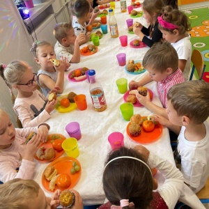 Dzieci siedzą przy stole w sali przedszkolnej, jedzą kolorowe owoce, piją sok.