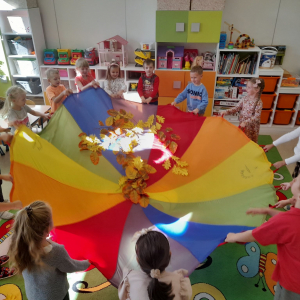 Dzieci bawią się w sali przedszkolnej chustą animacyjną, podrzucają do góry kolorowe liście.