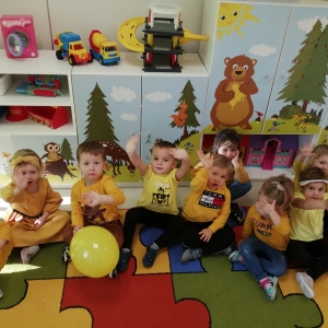 Na zdjęciu widac dzieci z grupy Biedronki, które siedzą na dywanie w sali przedszkolnej, trzymają w rękach żółty balonik.