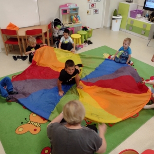 Dzieci siedzą na dywanie i bawią się kolorową chustą animacyjną. Jeden chłopiec siedzi na środku.