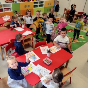 Dzieci siedzą przy stolikach w sali przedszkolnej i pokazują rysunki.