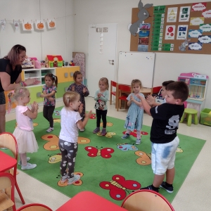 Dzieci bawią się w swojej sali. Stoją w kole na dywanie i bawią się z nauczycielem.

