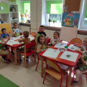 Dzieci siedzą przy stolikach w sali przedszkolnej i rysują. 