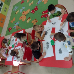 Dzieci siedzą przy stolikach w sali przedszkolnej i rysują.