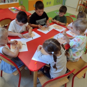 Dzieci siedzą przy stolikach w sali przedszkolnej i rysują. 