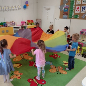 Dzieci bawią się w sali. Stoją w kole i trzymają w dłoniach kolorową chustę animacyjną.