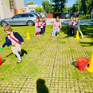 Dzieci bawią się na placu zabaw przy przedszkolu. Nauczyciel prowadzi z dziećmi zawody sportowe.
