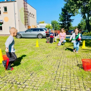 Dzieci bawią się na placu zabaw przy przedszkolu. Nauczyciel prowadzi z dziećmi zawody sportowe.