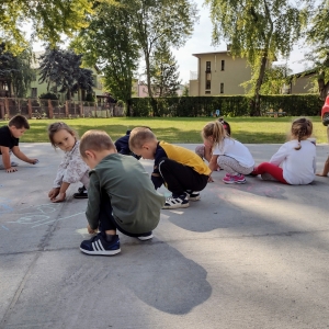 Dzieci bawią się na placu zabaw przy przedszkolu. Rysują kredą na betonie.