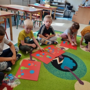 Dzieci siedzą na dywanie w swojej sali przedszkolnej. Dzieci bawią się kolorowymi kropkami.