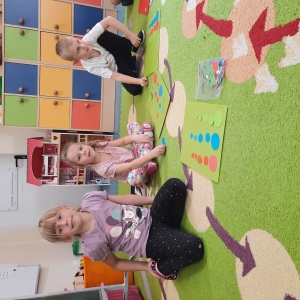 Dzieci siedzą na dywanie w swojej sali przedszkolnej. Dzieci bawią się kolorowymi kropkami.