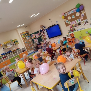 Dzieci siedzą przy stolikach w swojej sali. Bawią się kolorowymi balonami. Jest święto Przedszkolaka. Cieszą się.