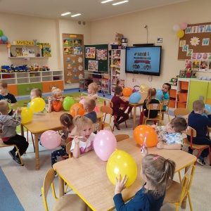 Dzieci siedzą przy stolikach w swojej sali. Bawią się balonami. Jest święto Przedszkolaka. Cieszą się.