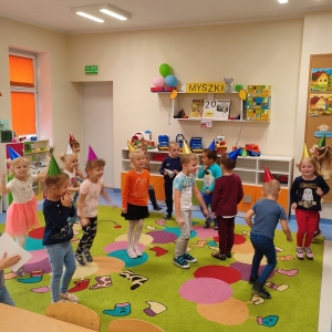 Dzieci tańczą na dywanie  w sali. Jest dzień przedszkolaka. Dzieci są radosne.