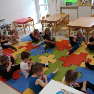 Dzieci siedzą na dywanie w swojej sali przedszkolnej. Dzieci ćwiczą.