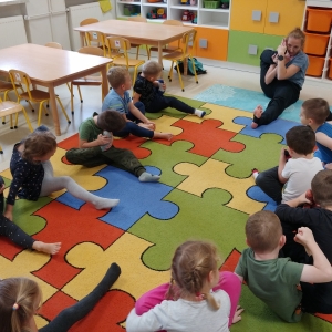 Dzieci siedzą na dywanie w swojej sali przedszkolnej. Dzieci ćwiczą.