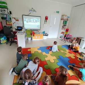 Dzieci siedzą na dywanie w swojej sali przedszkolnej. Dzieci oglądają film.