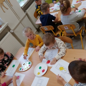 Dzieci siedzą przy stolikach i malują farbami na kartce.